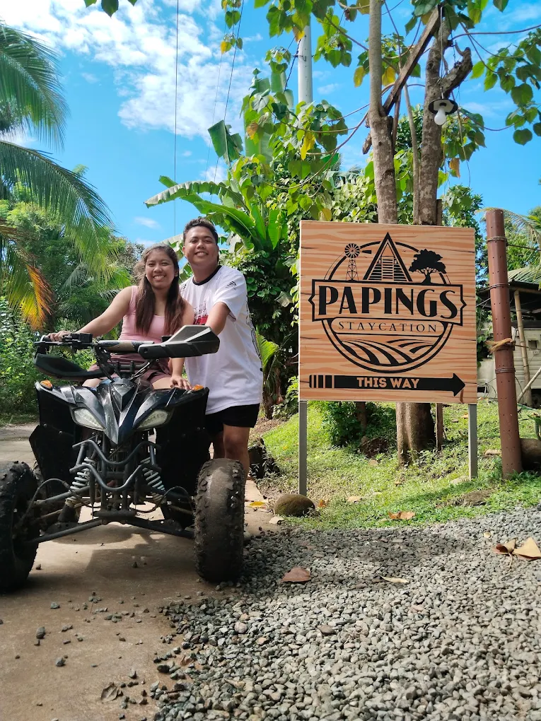 Couple on an ATV near Paping's Staycation and Campsite sign in Magallanes, surrounded by lush greenery.