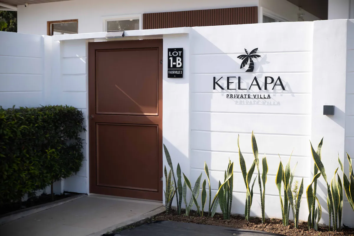 Entrance of Kelapa Private Villa in Alfonso, featuring a brown door, white wall, and decorative plants.