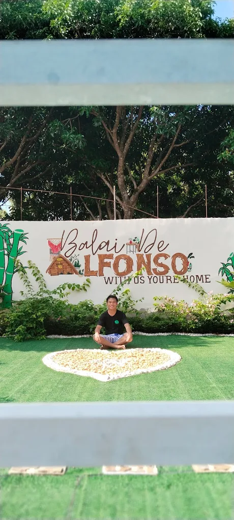 Person sitting on grass in front of Balai de Alfonso sign with trees in the background, Alfonso, Cavite.