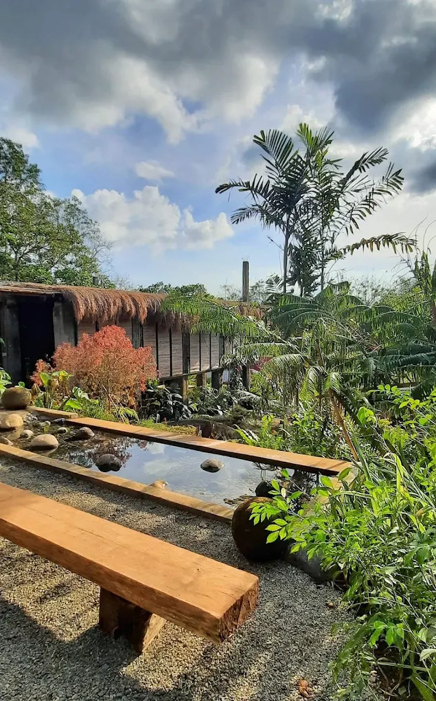 Tropical garden with wooden bench and pond at Maka Forest Villa, Alfonso, surrounded by lush greenery and cloudy sky.