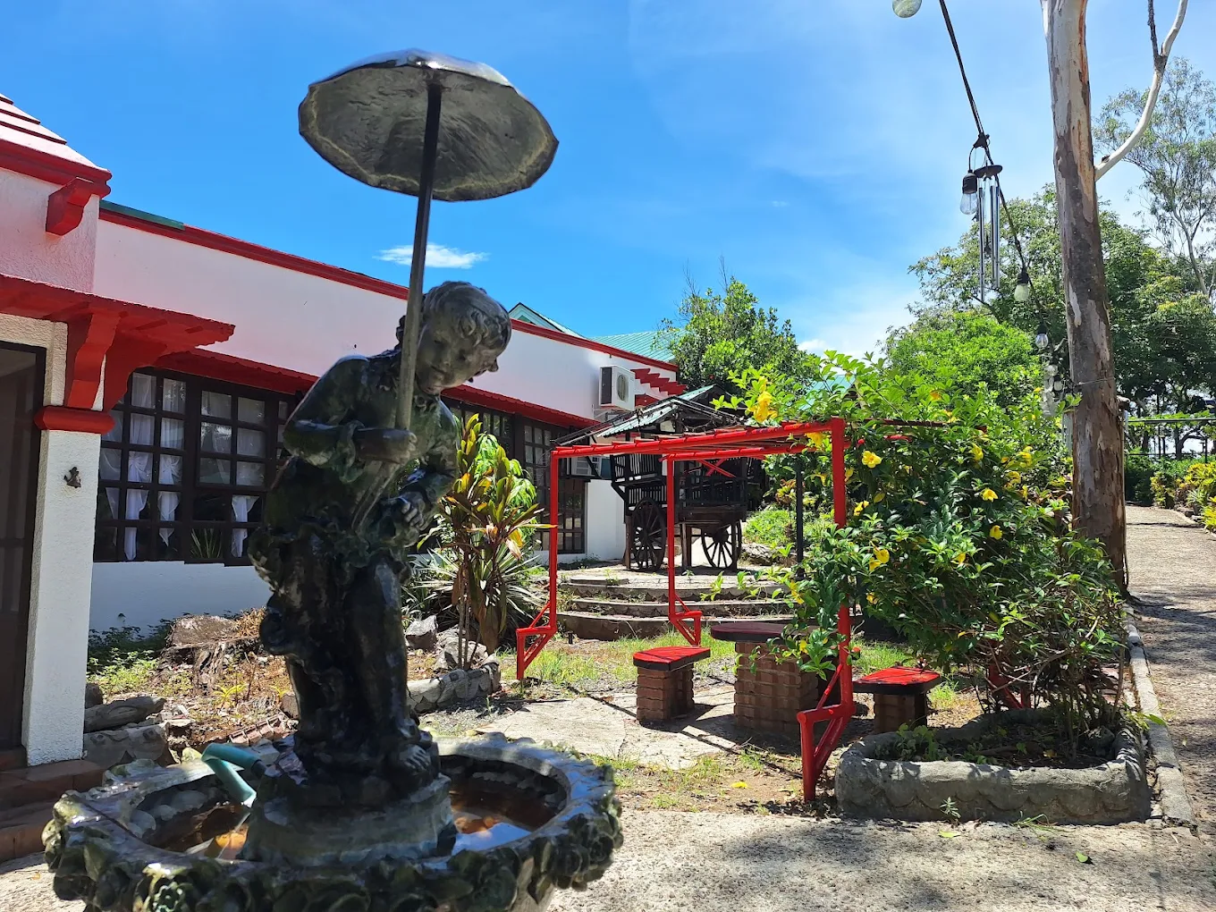 Fountain statue and garden seating at Balay Indang, Indang, with red-roofed building and lush greenery in the background.