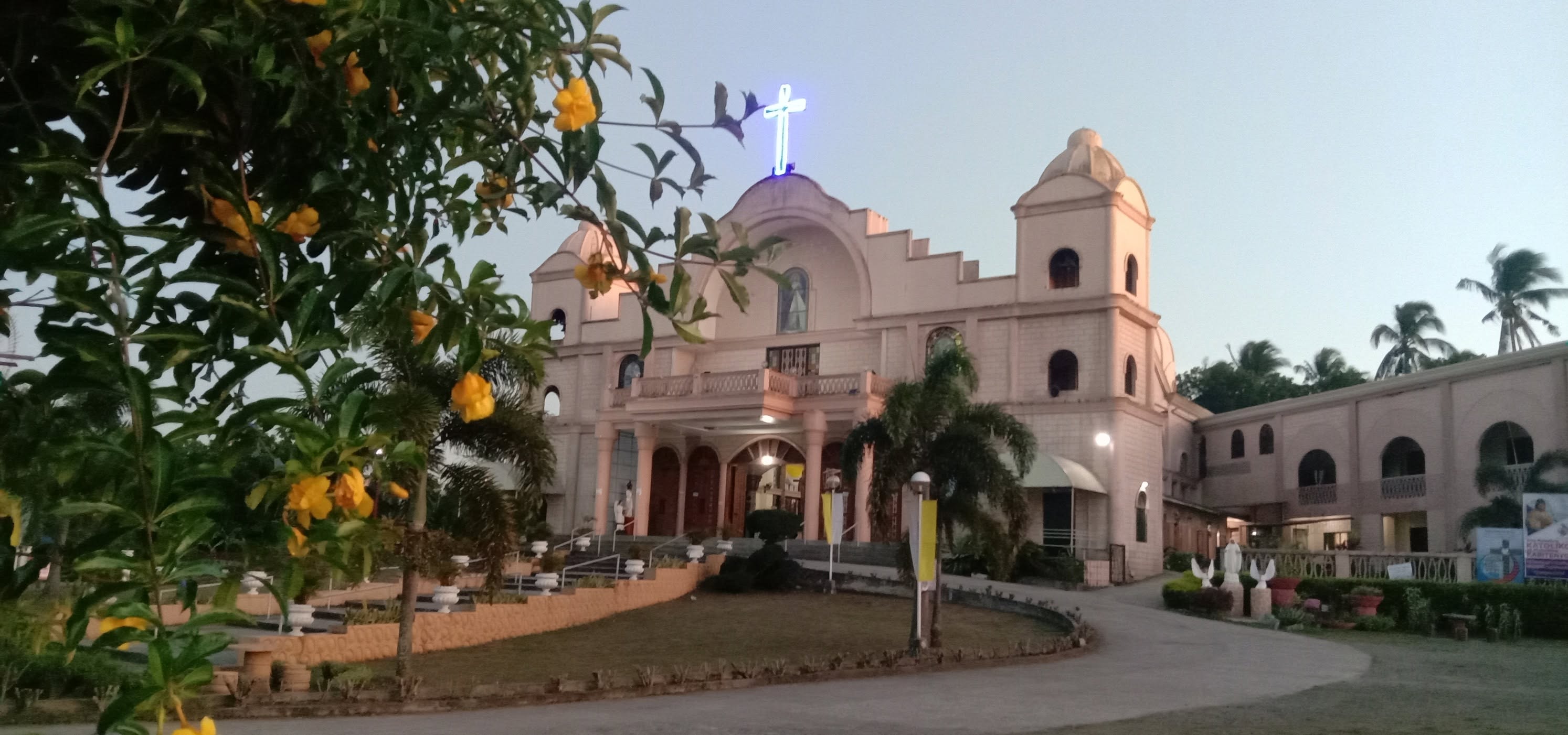 Divine Mercy Parish in Silang with a lit cross, surrounded by palm trees and yellow flowers at dusk.