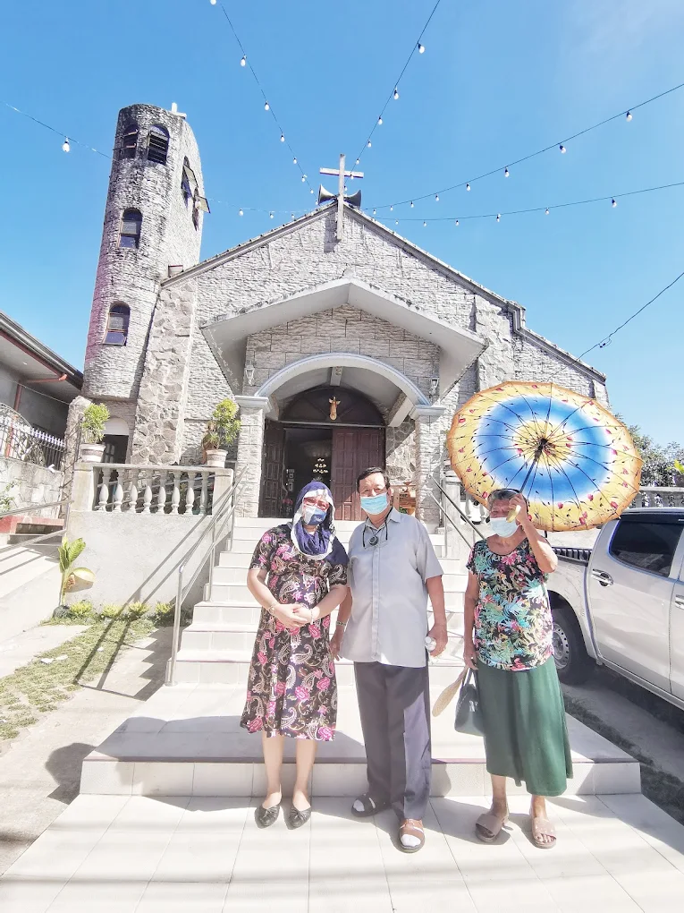 Stone facade of San Juan Bautista Parish in Silang with three people standing in front, one holding a colorful umbrella.